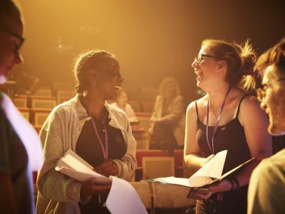 Two people standing up wearing lanyards in a theatre with clipboards and paper. They are smiling at each other.